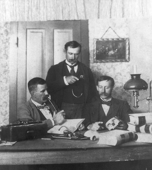 Three white men with mustaches and suits before a desk, one holding part of a human skull