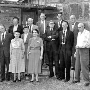 Group of older white men in suits and women in dresses standing outside brick building