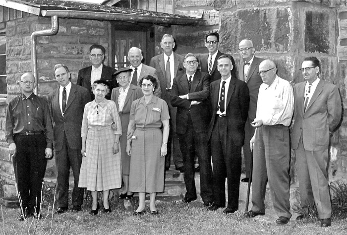 Group of older white men in suits and women in dresses standing outside brick building