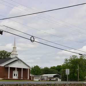 Single-story brick church building with steeple and covered entrance on parking lot with brick sign