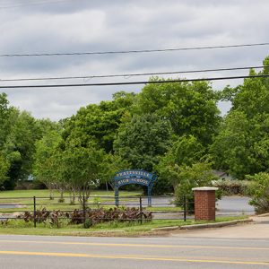 Metal fence with brick columns and parking lot with blue metal sign and trees on street