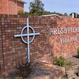 Brick "Presbyterian Village" sign with cross logo and flower bed on parking lot