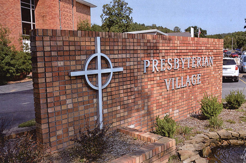 Brick "Presbyterian Village" sign with cross logo and flower bed on parking lot