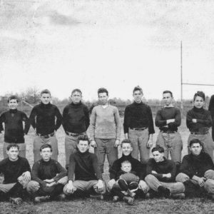Group of young white men in casual clothing and baby boy, along with one older white man in suit, posing on football field