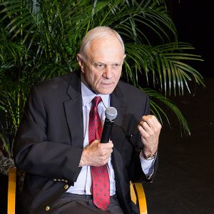 Old white man in suit and tie with microphone sitting on stage