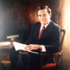 White man in suit and tie sitting at his desk and posing with an open book