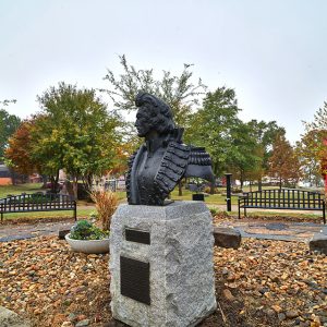 Bust of man in military uniform on pedestal in park flower bed with rocks and benches in the background