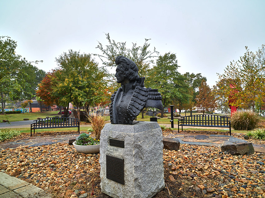 Bust of man in military uniform on pedestal in park flower bed with rocks and benches in the background