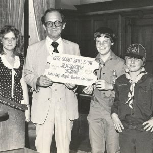 White man with glasses in suit  and white woman standing with white boys in scout uniforms holding a giant ticket
