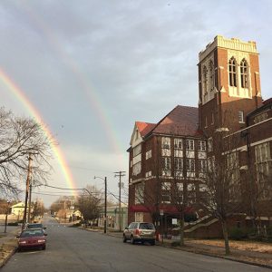 Rainbow over city street with parked cars and multistory brick church with tower on the right
