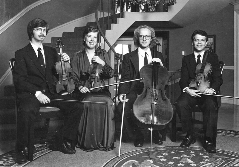 Three white men, one white woman, seated, holding stringed instruments before staircase