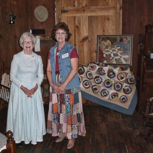 Two white women in dresses inside log cabin with quilt displayed behind them