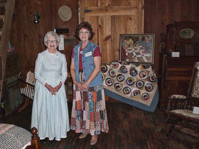 Two white women in dresses inside log cabin with quilt displayed behind them