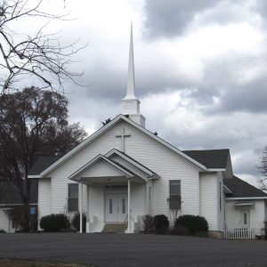 Church building with cross above covered entrance and steeple with parking lot in front