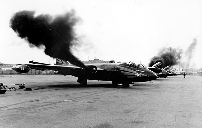 row of airplanes with first plane having dark smoke coming from its engines and more smoke coming from one of the other planes