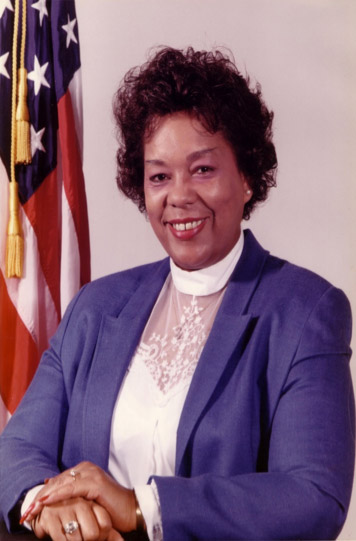 African-American woman smiling in purple suit and white shirt with American flag behind her