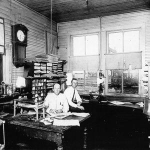 Two white men in office with desks shelves and clock