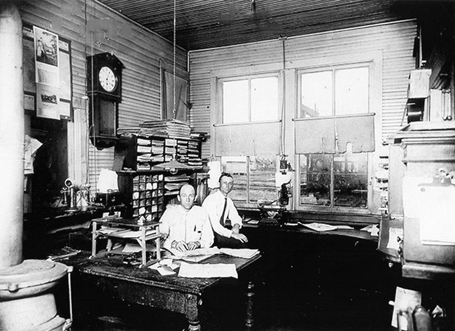 Two white men in office with desks shelves and clock