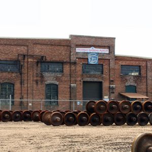Rows of train wheels and axles piled on top of each other inside fence outside multistory brick building