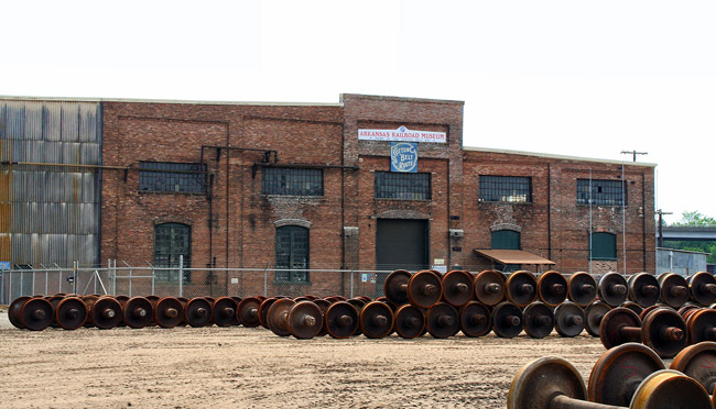 Rows of train wheels and axles piled on top of each other inside fence outside multistory brick building