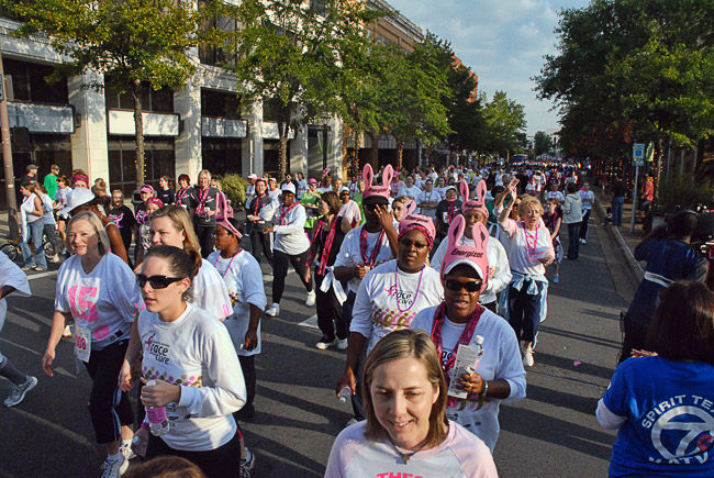 Crowd of white and African-American women marching on city street wearing white shirts with pink accouterments