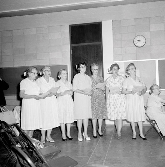 Group of older white women standing in a row holding sheet music in dresses and heels with bows in their hair