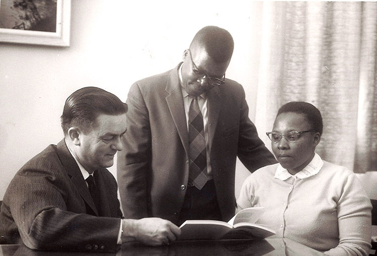 White man in suit sitting at table reading to African-American man standing and African-American woman sitting beside him