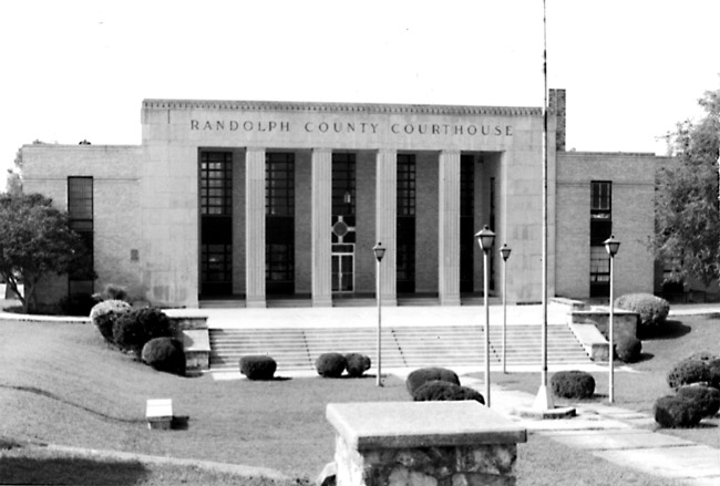 Multistory stone brick building with carved "Randolph County Courthouse" art deco columns and large courtyard with round bushes