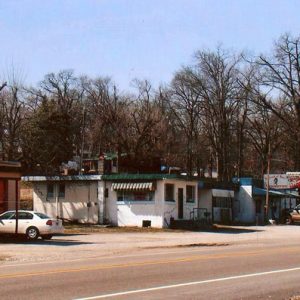 Single-story stores and two-car garage building on two-lane road with vehicles parked nearby