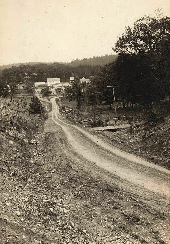 Looking down dirt road on hill with multistory buildings in the background and trees on the right