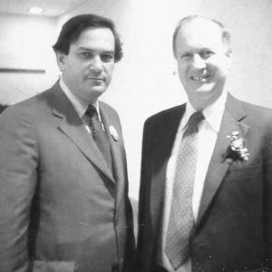 Two white men in suits and ties with one wearing a button and one wearing a corsage