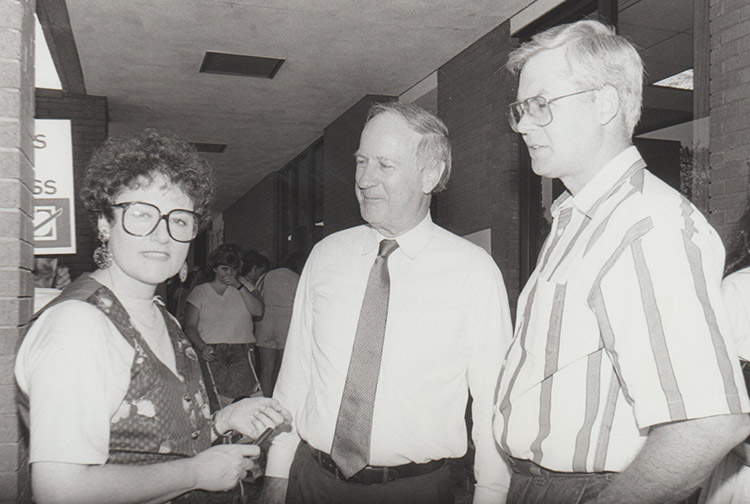 Pair of older white men speaking to white woman with glasses and vest in hallway