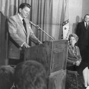 White man in suit speaking at lectern with wife and white crowd around him