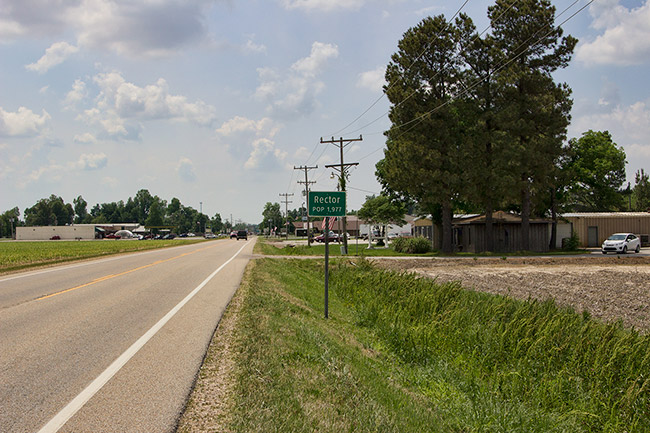 Two-lane road with "Rector" sign and single-story buildings in the background