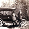Two-white men in military uniform with car featuring the Red Cross emblem