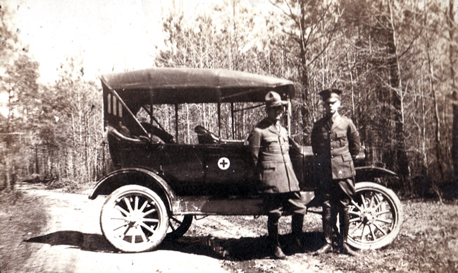 Two-white men in military uniform with car featuring the Red Cross emblem