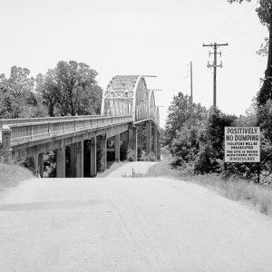 Steel arch bridge with concrete platform over river with sign "Positively No Dumping" to the right