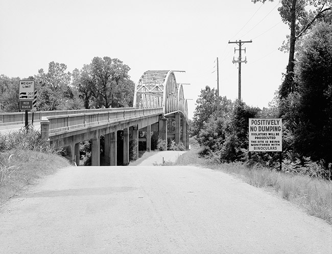 Steel arch bridge with concrete platform over river with sign "Positively No Dumping" to the right