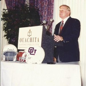 White man in suit and tie speaking at lectern with football helmets on a table nearby