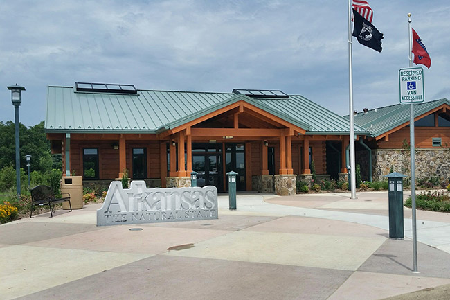 Log cabin building with green roof flags and "Arkansas the natural state" sign on concrete