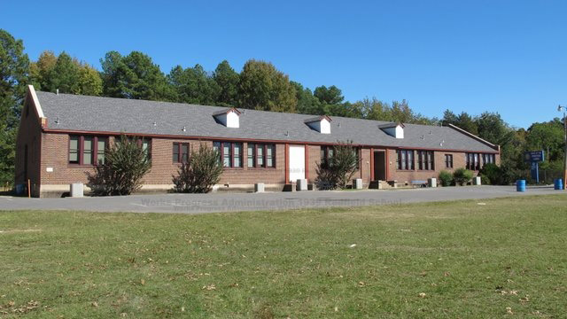 Single-story brick school building with rows of windows and parking lot with sign