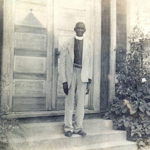 Older African-American man in suit with white clerical collar standing outside church doors next to a large plant