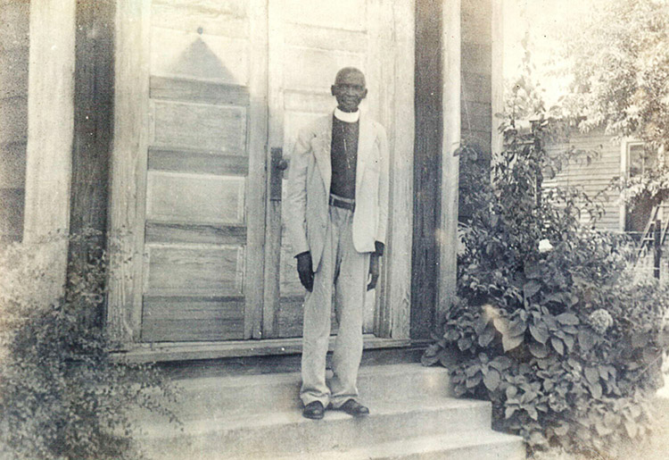 Older African-American man in suit with white clerical collar standing outside church doors next to a large plant