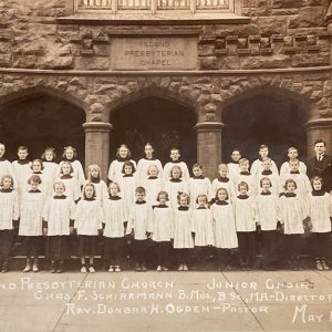 White man in suit and tie standing with group of white children and white man in clerical robes outside multistory church building
