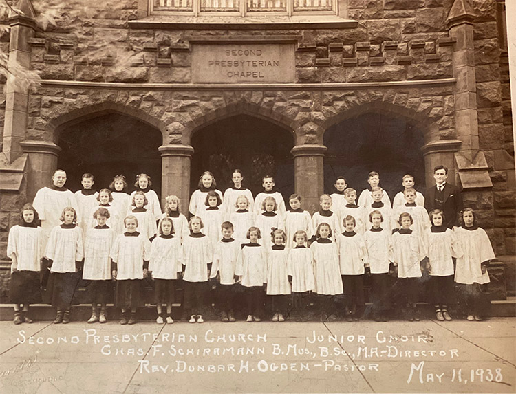 White man in suit and tie standing with group of white children and white man in clerical robes outside multistory church building