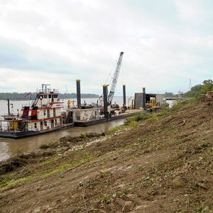 Tugboat pushing construction barge with crane on river and bulldozers on shore