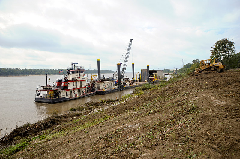 Tugboat pushing construction barge with crane on river and bulldozers on shore