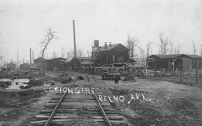 Railroad tracks with industrial buildings and water tower signed "cotton gins Reyno Ark"