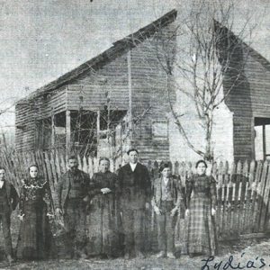 White men and women  with dog posing in a line outside house with brick chimney and fence