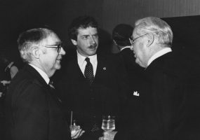 White man with mustache in suit and tie standing in between two older white men in suits with glasses talking
